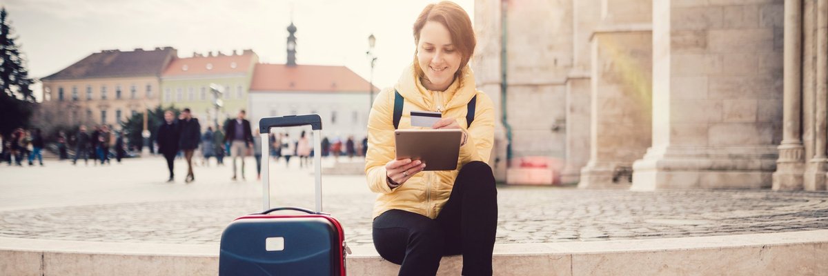 A woman sitting in a sunny plaza next to her suitcase while she looks something up on her tablet and holds her credit card.
