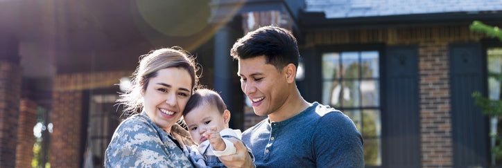 A young military veteran standing in front of her home with her baby and husband.