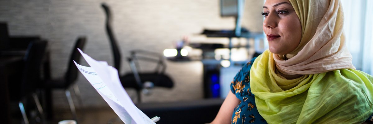 A person wearing a headscarf sitting at a desk with a laptop and holding papers.