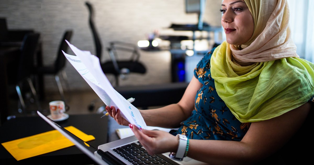 A person wearing a headscarf sitting at a desk with a laptop and holding papers.