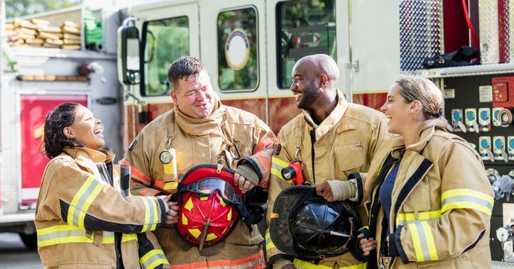 Four firefighters talking and laughing in their gear while standing beside a fire truck.