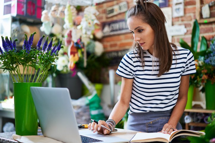 An owner of a flower shop working on a laptop at the counter while writing in a notebook.