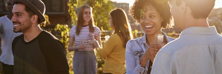 A group of smiling young adults drinking cocktails at a rooftop party at sunset.