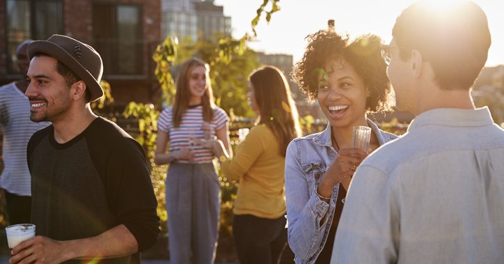 A group of smiling young adults drinking cocktails at a rooftop party at sunset.