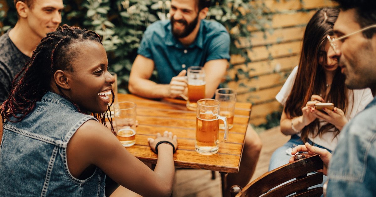 A group of laughing friends drinking beers at a restaurant patio table.