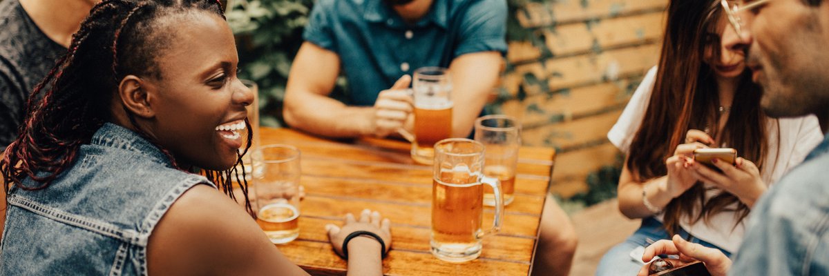A group of laughing friends drinking beers at a restaurant patio table.