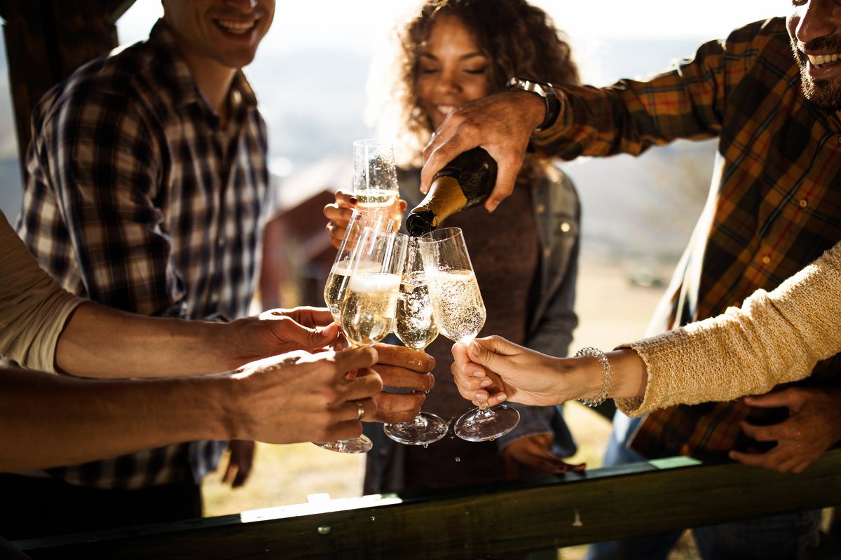 A group of friends holding up champagne glasses to cheers while someone pours a bottle.