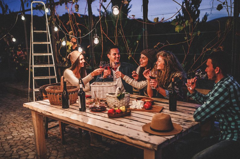 A group of young adult friends drinking wine at a picnic table in a decorated backyard patio.