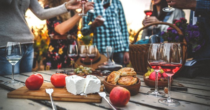 A group of friends drinking wine around a cheese plate on a picnic table in a vineyard.