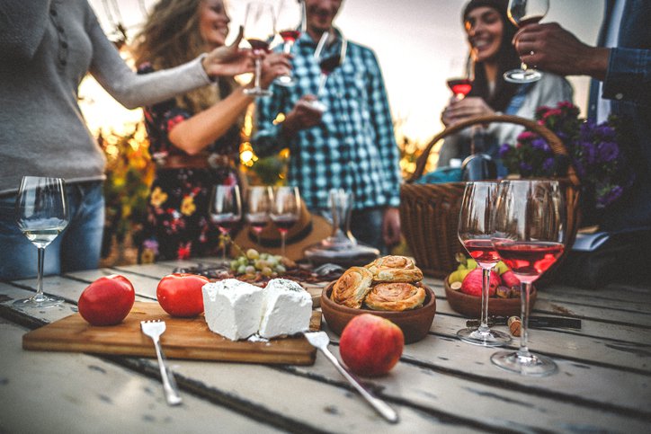 A group of friends drinking wine around a cheese plate on a picnic table in a vineyard.