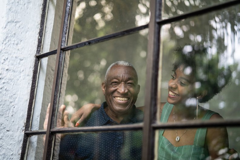 A smiling grandfather and his adult granddaughter looking out of window at the trees.