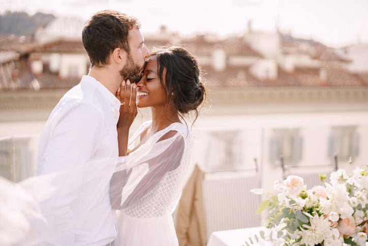 A bride and groom on a sunny rooftop while he kisses her forehead and she smiles.