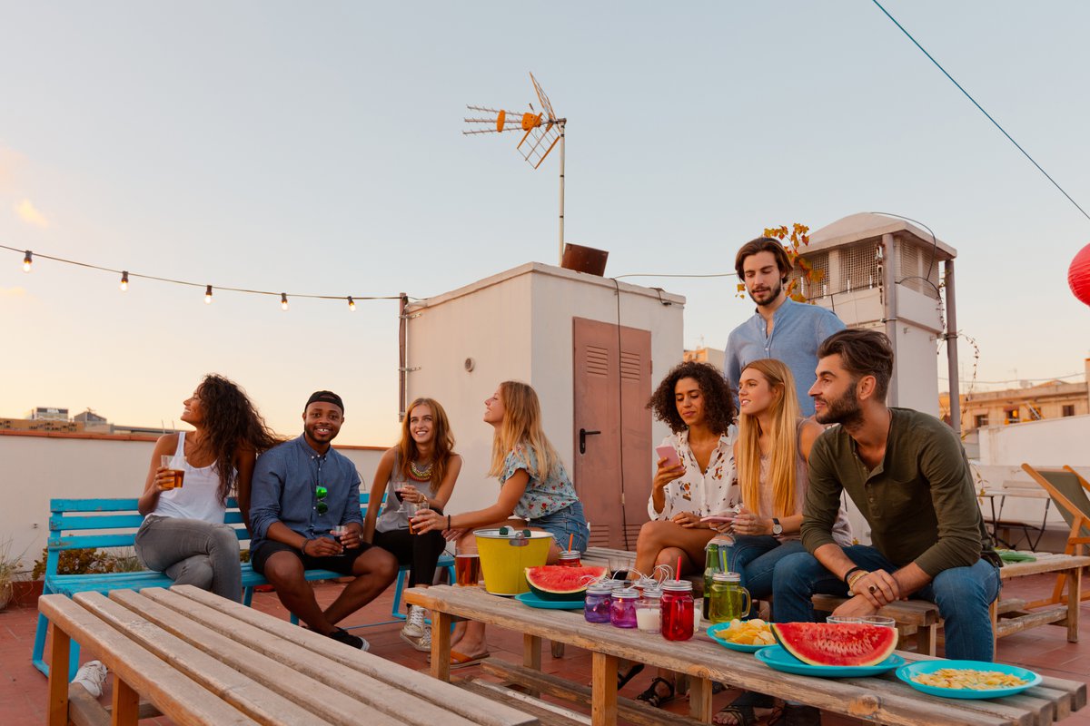 A group of young adult friends hanging out on a decorated rooftop with cocktails at dusk.