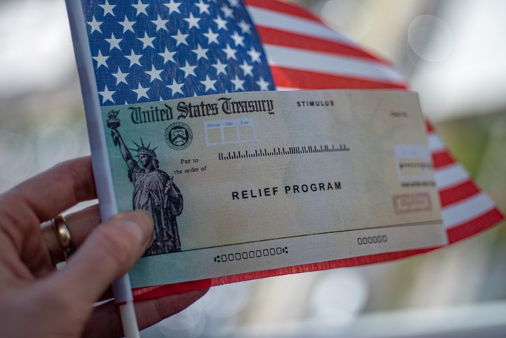 A hand holding a small American flag and a stimulus check from the U.S. Treasury.