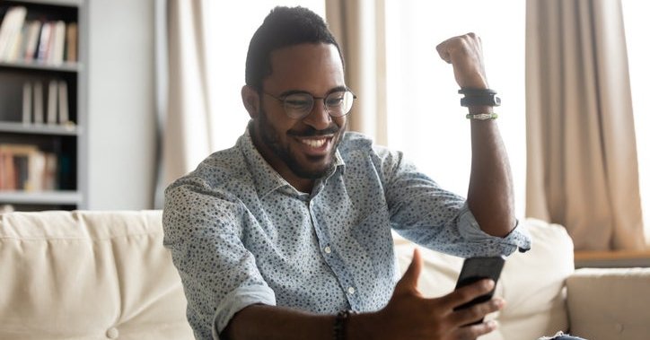 A man pumping his fist in excitement while sitting on his couch and looking at something on his phone.