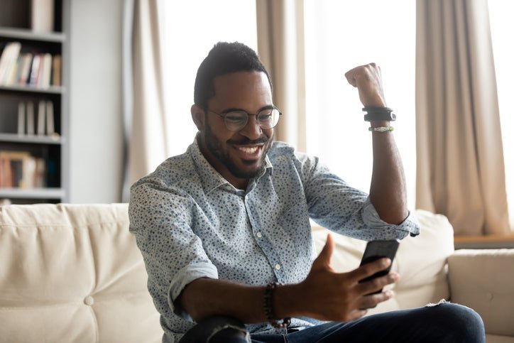 A man pumping his fist in excitement while sitting on his couch and looking at something on his phone.