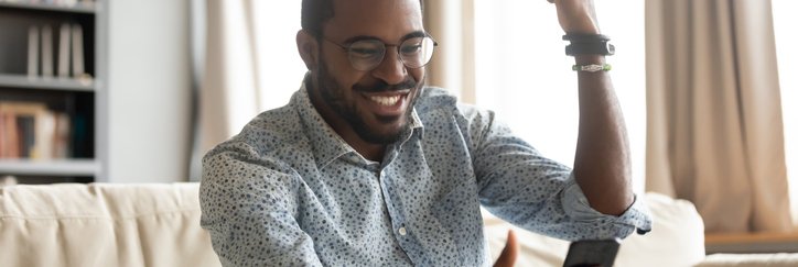 A man pumping his fist in excitement while sitting on his couch and looking at something on his phone.