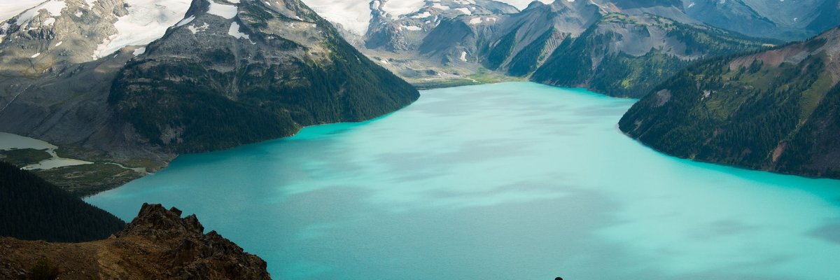 Two hikers holding hands and standing on a lookout point over a glacial lake surrounded by snow-capped mountains.