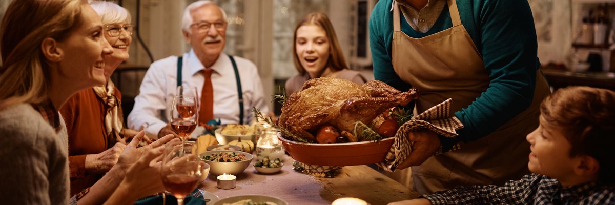 Group of smiling adults sitting around a Thanksgiving table full of food in a dining room decorated with twinkle lights while one person sets the cooked turkey down.