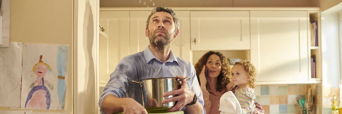 Man standing below leaky ceiling catching water in a bucket.