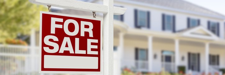 A For Sale sign in front of a sunny green lawn and white two-story home.