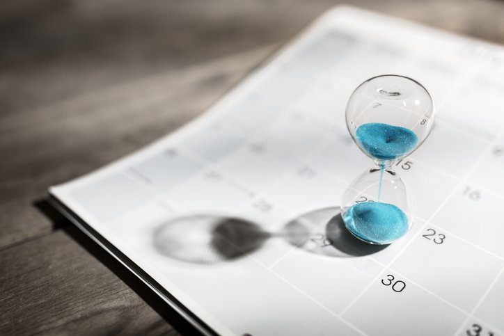 An hourglass filled with blue sand sitting on top of a calendar page on a wooden desk.