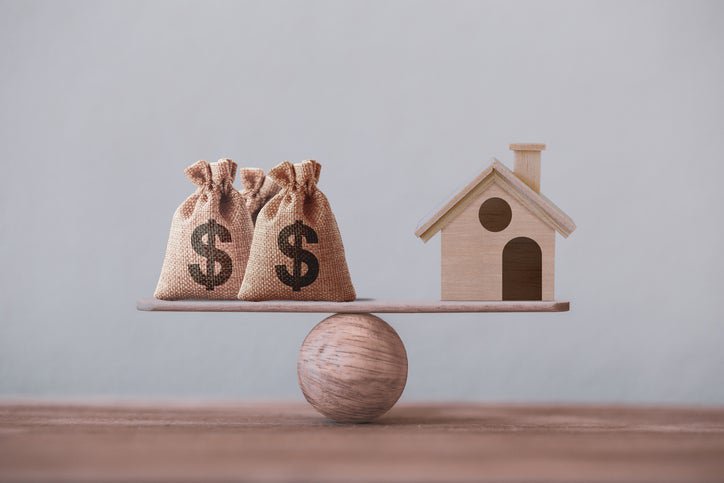 A toy house and bags of money balanced on a plank resting on a wooden ball.