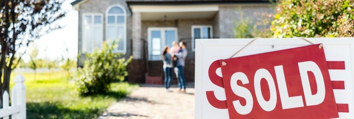 A Sold sign in front of a driveway leading up to a family standing in front of their new home.