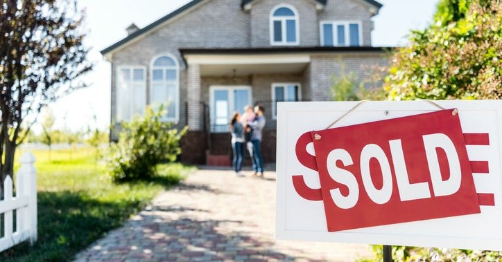 A Sold sign in front of a driveway leading up to a family standing in front of their new home.