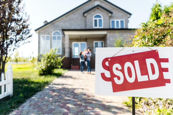 A Sold sign in front of a driveway leading up to a family standing in front of their new home.