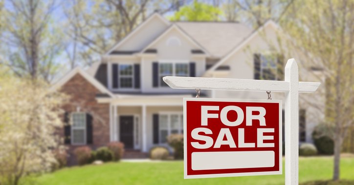 A For Sale sign in front of a two-story house surrounded by trees and a lawn.