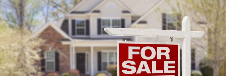 A For Sale sign in front of a two-story house surrounded by trees and a lawn.