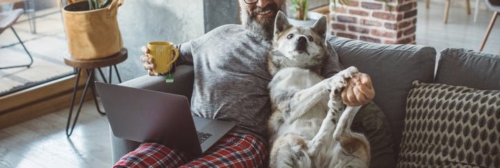 A man sitting on the couch with a laptop and cup of coffee and playing with his dog next to him.