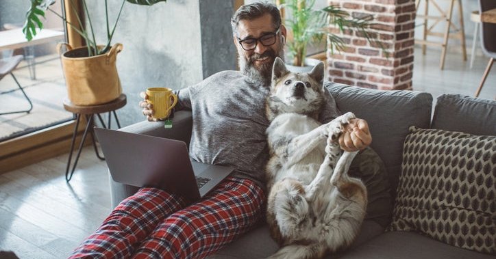 A man sitting on the couch with a laptop and cup of coffee and playing with his dog next to him.
