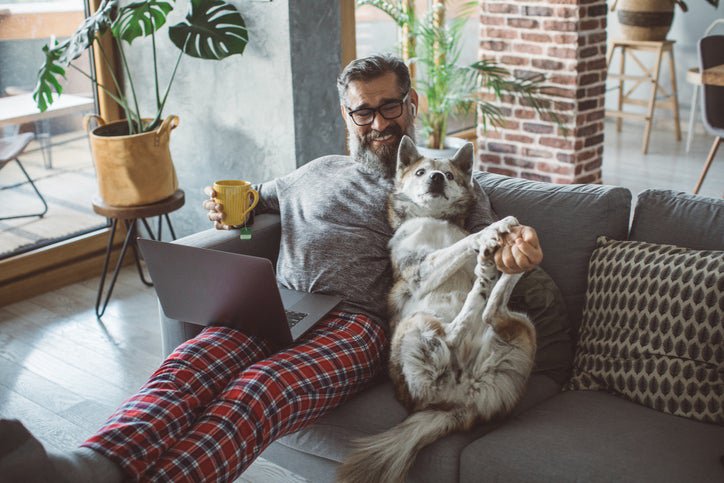A man sitting on the couch with a laptop and cup of coffee and playing with his dog next to him.