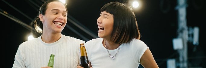A laughing man and woman drinking beer while standing on a rooftop under string lights.
