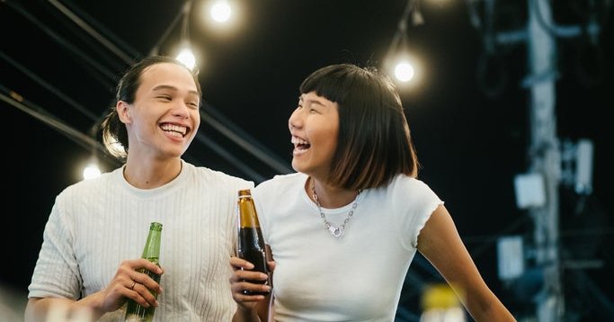 A laughing man and woman drinking beer while standing on a rooftop under string lights.