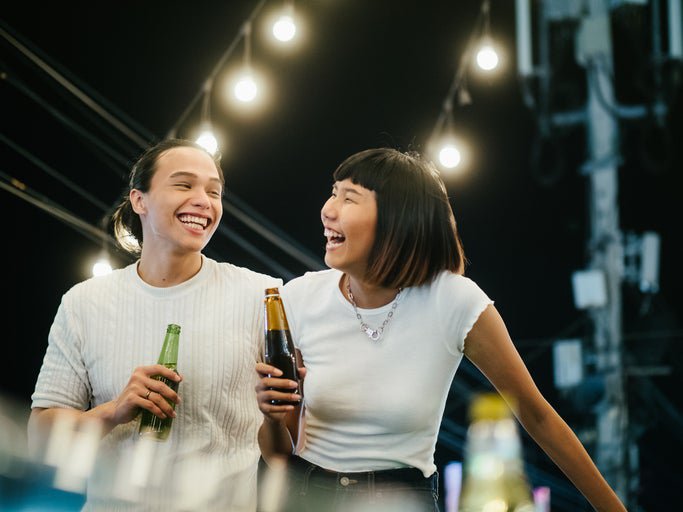 A laughing man and woman drinking beer while standing on a rooftop under string lights.