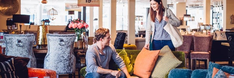 A man and woman looking at furniture in a department store with sales signs hanging from the ceiling.
