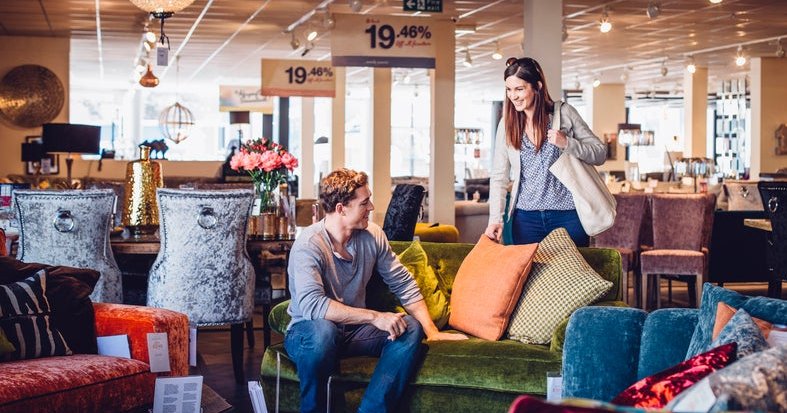 A man and woman looking at furniture in a department store with sales signs hanging from the ceiling.