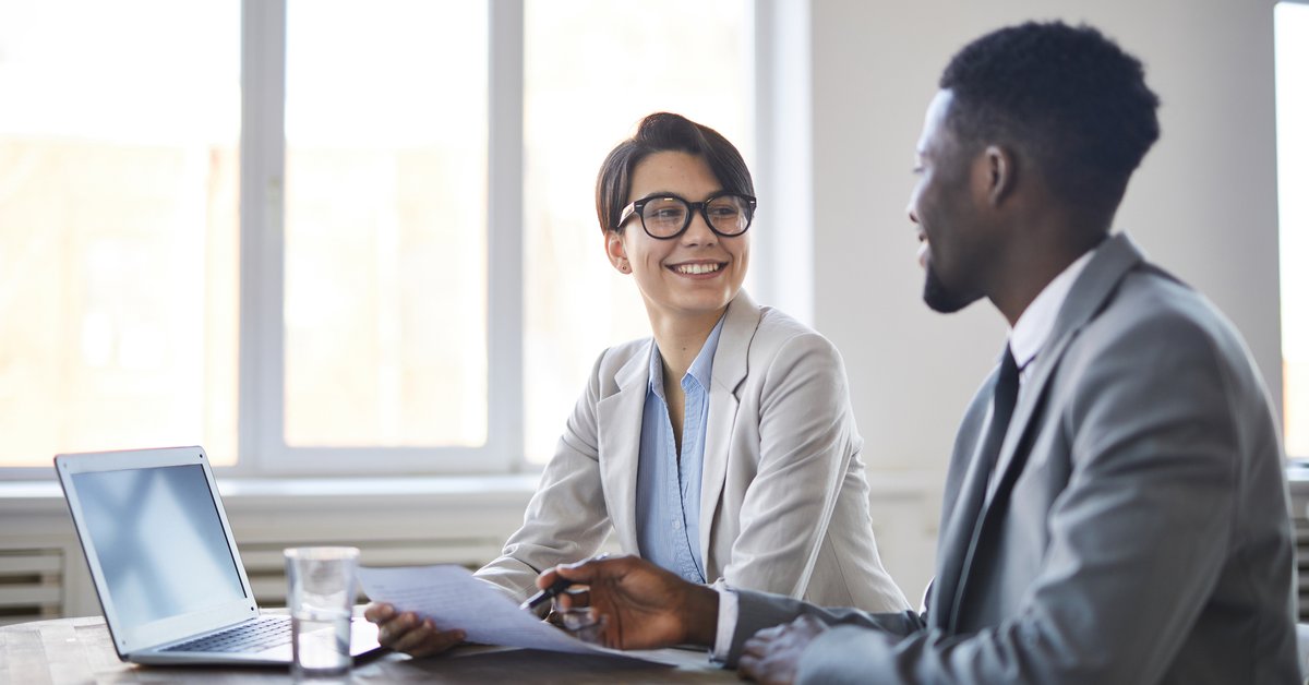 man and woman in business attire sitting at table and talking over documents.