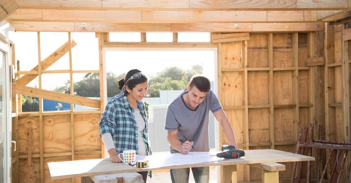 A man and woman standing in a partially constructed new home and looking at plans on a work table.