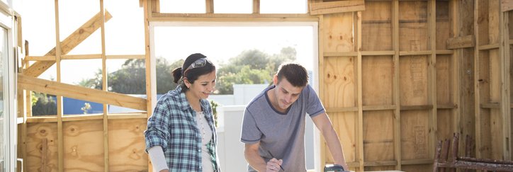 A man and woman standing in a partially constructed new home and looking at plans on a work table.