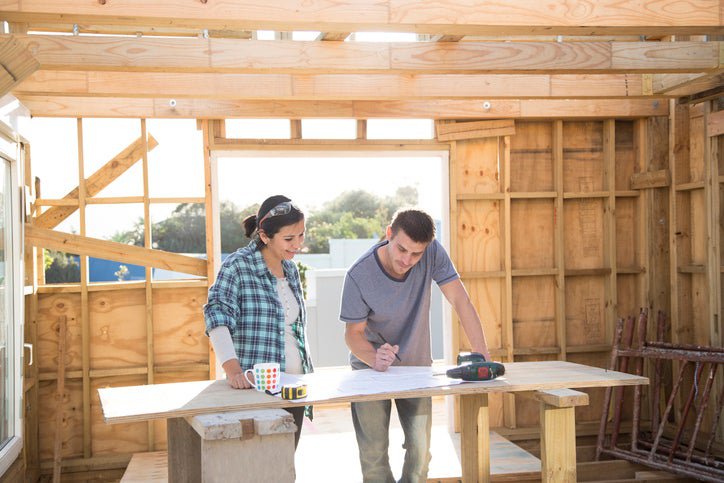 A man and woman standing in a partially constructed new home and looking at plans on a work table.