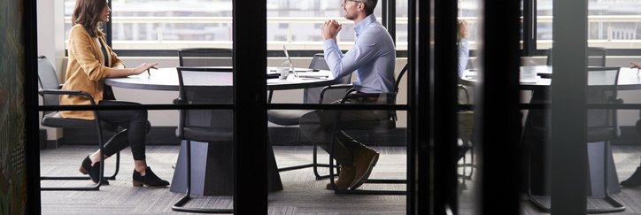 A man and woman sitting at opposite ends of a table in an office meeting room for a job interview.