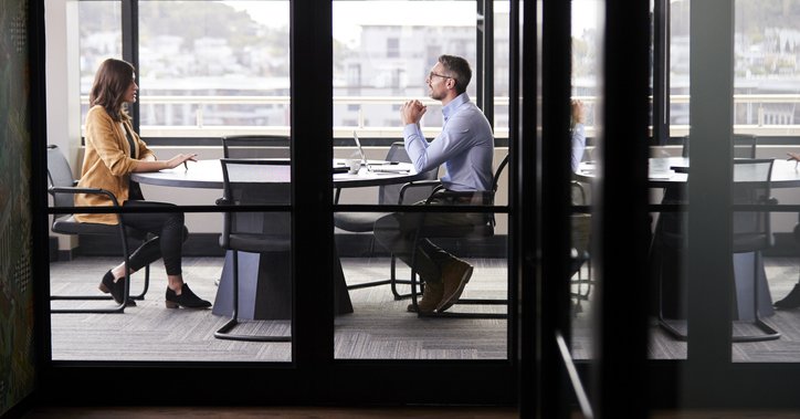 A man and woman sitting at opposite ends of a table in an office meeting room for a job interview.