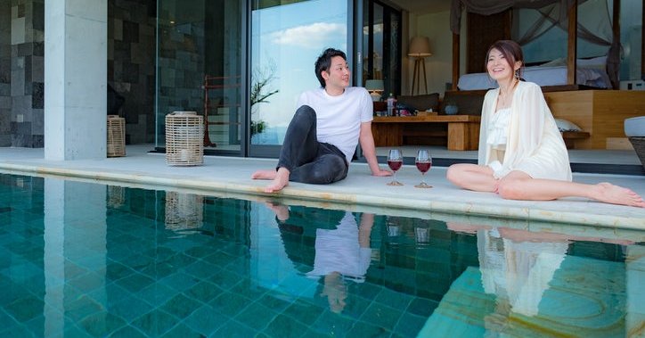 A man and woman sitting at the edge of a pool outside their room in a luxury hotel.