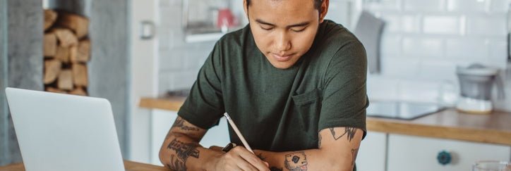 A man sitting at his kitchen table in front of an open laptop and writing in a notebook.