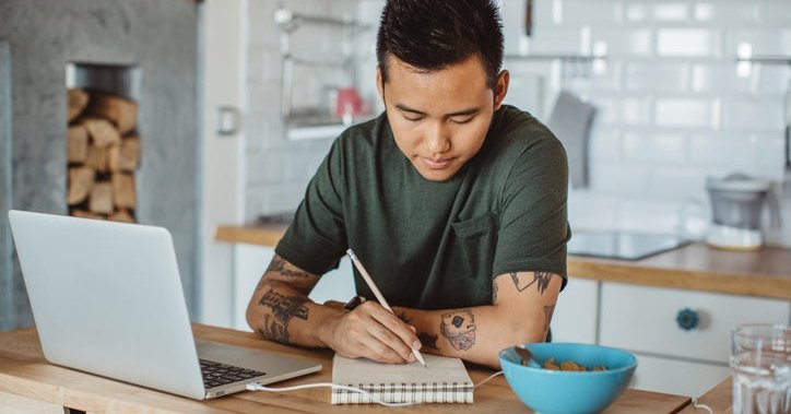 A man sitting at his kitchen table in front of an open laptop and writing in a notebook.