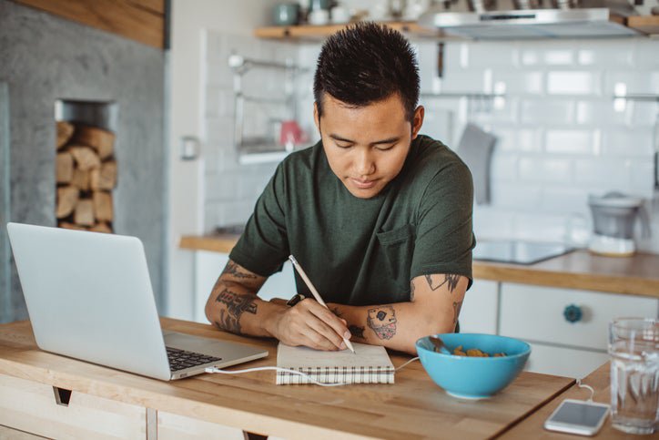 A man sitting at his kitchen table in front of an open laptop and writing in a notebook.